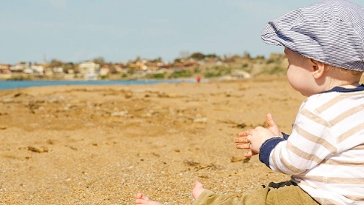 Clothed male baby wearing a flat cap sat on a beach with sand in his hands