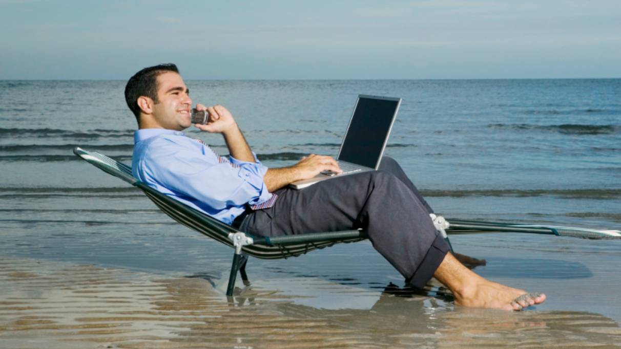 man wearing trousers and a button down shirt with his laptop and pone lounging on a beach