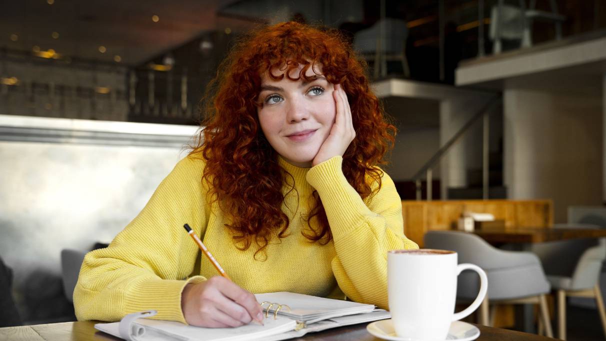 A person writing in a notebook with a warm drink on the table beside them. 