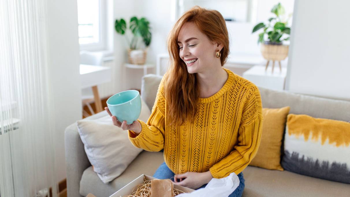 A person sitting on a sofa at home, unpacking a light blue mug from a delivery box