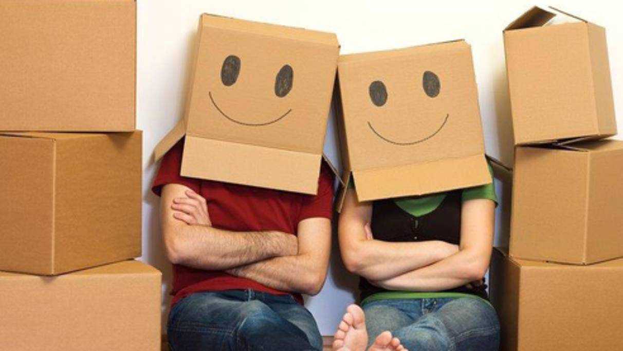 Couple sat between stacked boxes, both wearing boxes on their heads with smiley faces drawn on them