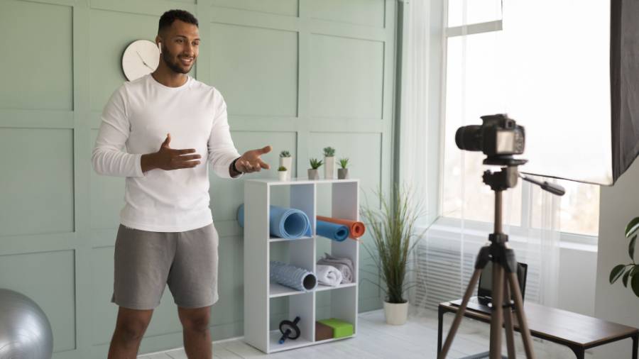 A person filming a video standing in front of a camera with fitness equipment in the background.