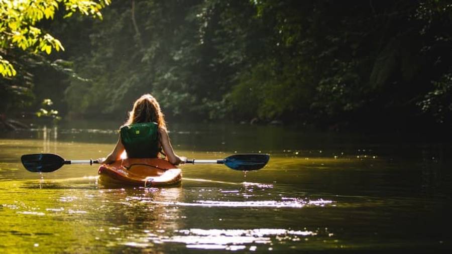 Woman wearing green backpack in kayak holding paddle, floating in green waters surrounded by greenery
