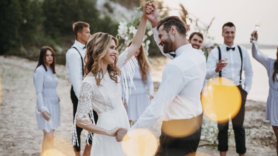 Couple dancing on a beach at sunset with guests in formal attire holding drinks