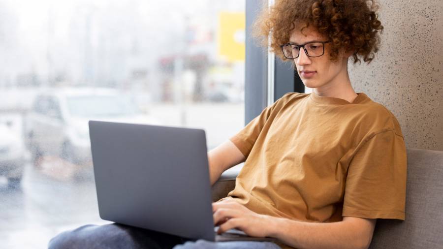 Someone in a brown T-shirt and jeans is sitting on a couch using a laptop, with large windows behind them showing buildings and cars outside