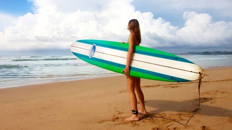 Female surfer standing on a beach, holding a surfboard, looking out to sea