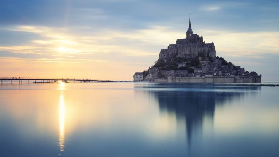 Rooves and spires of Mont Saint-Michel on a rocky island surrounded by water. Sunset reflects in the water