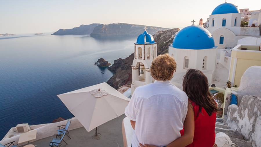 Couple sat embraced looking over the white and blue buildings of Greece along a rocky coastline 