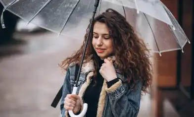 Person holding a transparent umbrella outdoors, wearing a denim jacket. The background shows a wet pavement and buildings