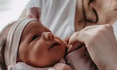 Baby girl in a pink outfit and matching headband, looking at her mum, holding her mum's hand