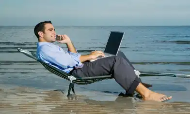 man wearing trousers and a button down shirt with his laptop and pone lounging on a beach