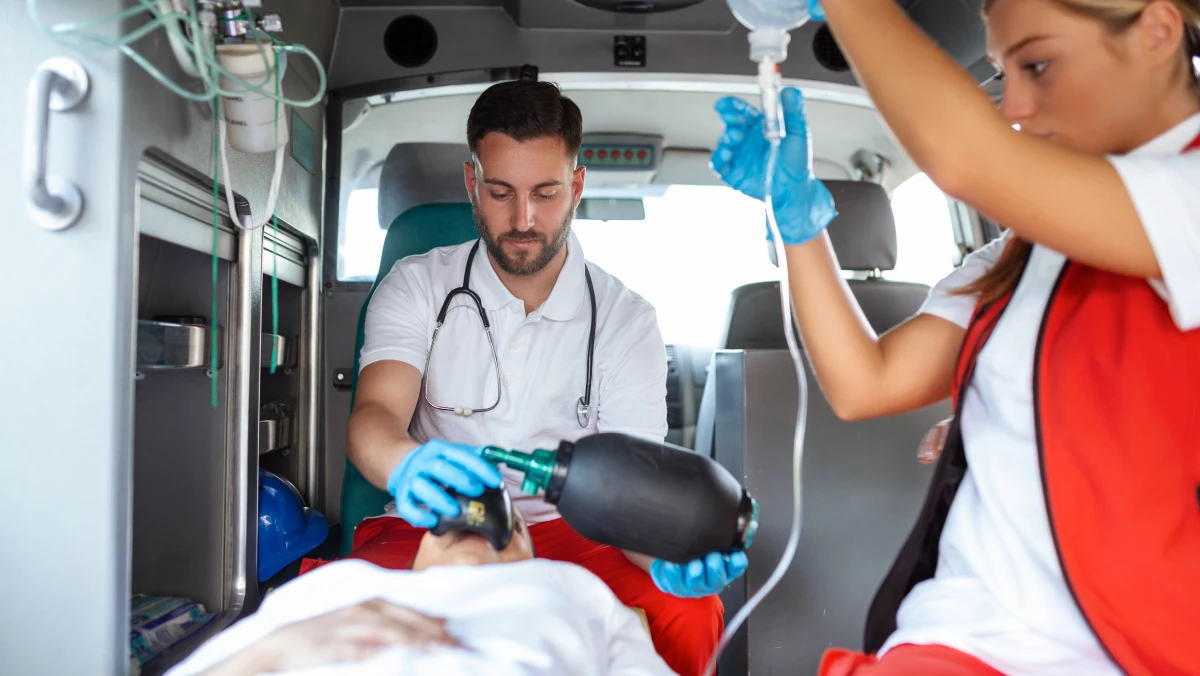 Two medical professionals in an ambulance attend to a patient on a stretcher, using a resuscitator and holding an IV bag