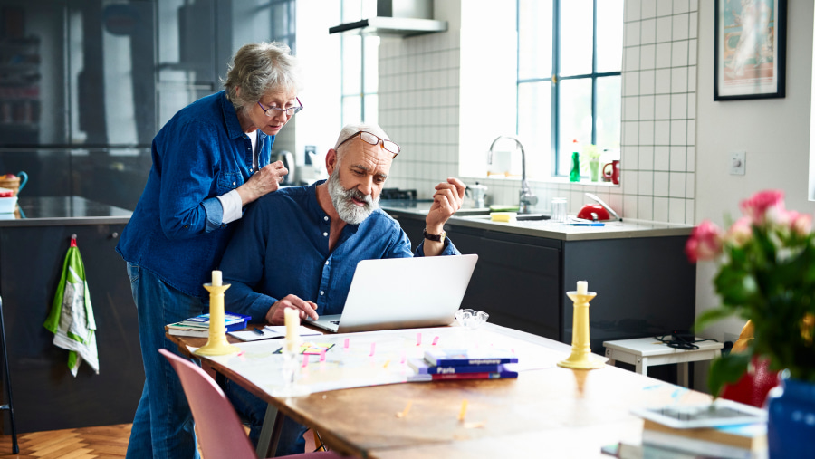 Elderly couple at the kitchen table with the laptop open, both looking at the screen 