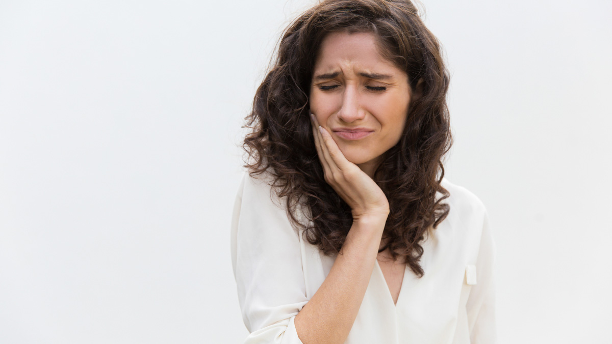 A woman with long brunette hair in a white shirt holds the side of her jaw with an expression of discomfort on her face 