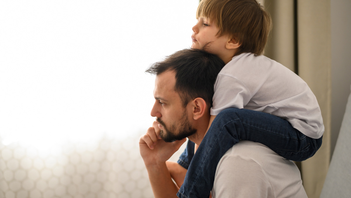 A man carries a young boy on his shoulders inside a home, his head in his hand and looking thoughtful 