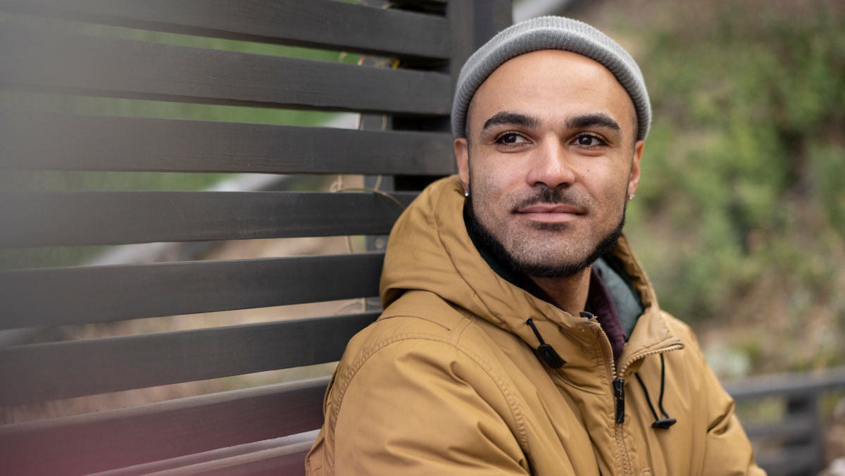 A man stands by a garden fence panel looking into the distance. He wears a grey beanie hat and a light brown or beige hooded jacket