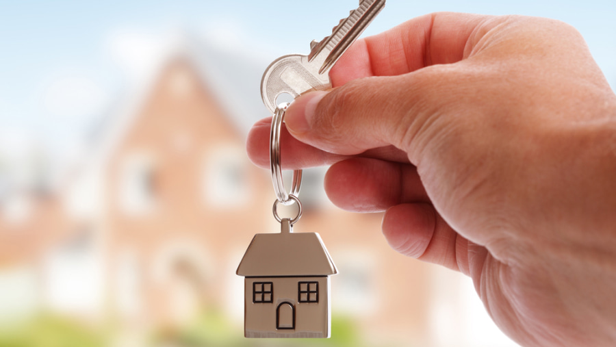 Hand holding up house keys with a house keyring attached to them, blurred houses in the background