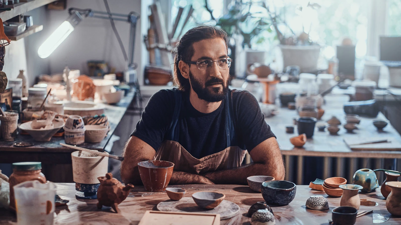 A man sitting at a workbench in a pottery studio with various pottery tools, brushes, and several pieces of pottery in different stages of completion