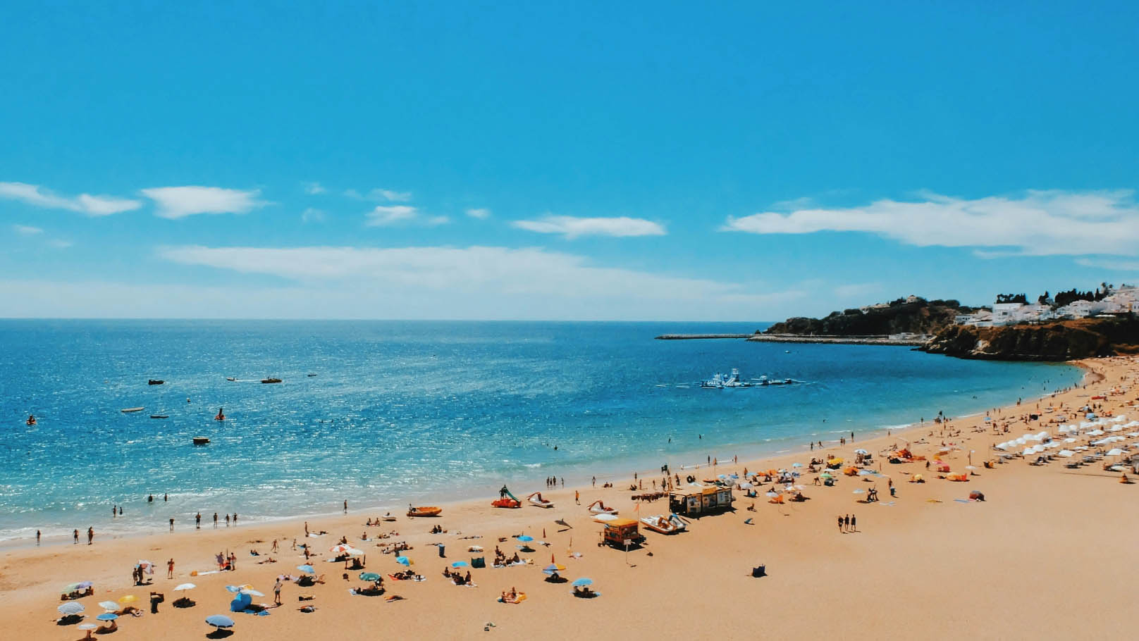 Raised view of a sandy beach where a light sea and sky. The beach is dotted with umbrellas, blankets and people enjoying their holidays. In the distance, small white buildings and dark vegetation line the shore.